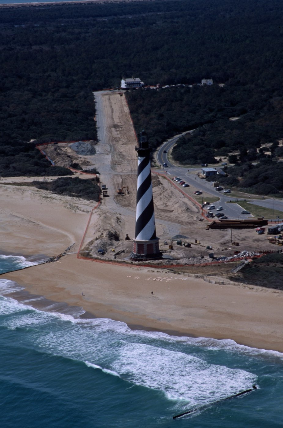 This aerial photograph shows how close the Cape Hatteras Lighthouse stood to the sea before its 1999 move; during strong storms, sea water washed around its base. The cleared move path is in the background