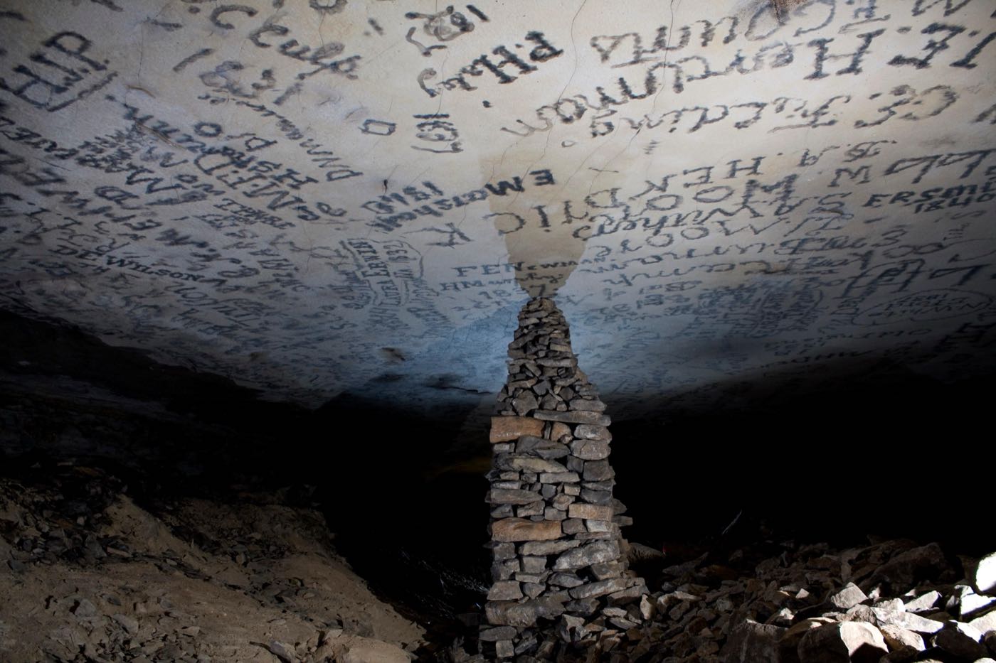 A large stacked stone pillar reaching a flat ceiling covered with signatures, in a Mammoth Cave passage